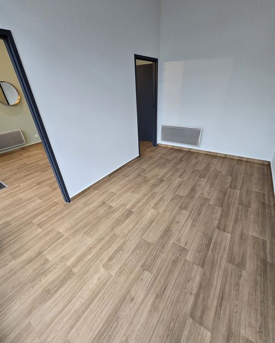 Pièce vide avec un sol imitation bois, des murs blancs et une porte donnant sur un couloir.