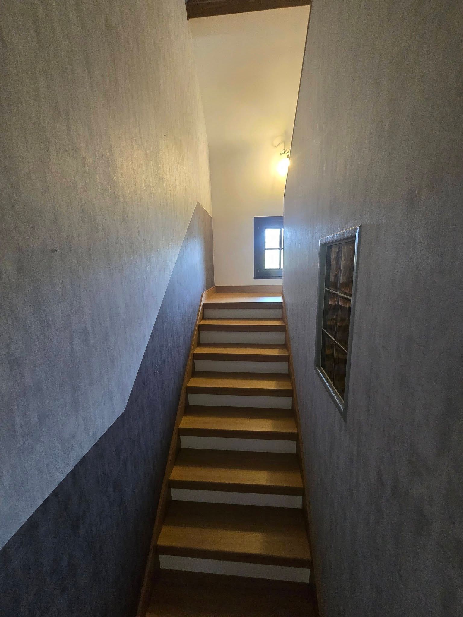 Escalier intérieur étroit aux murs beiges, avec une rampe sombre et une petite fenêtre en haut.