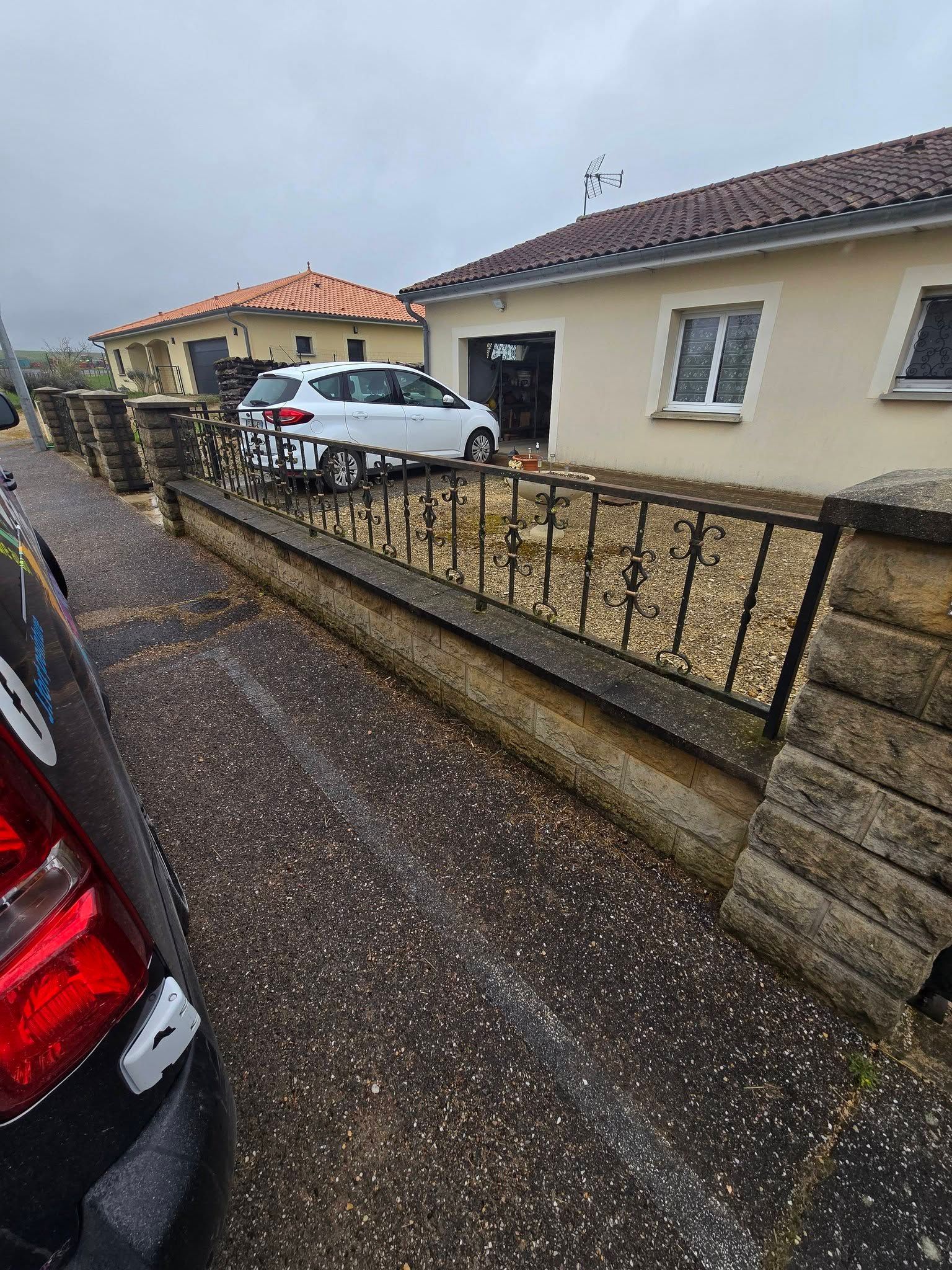 Allée résidentielle avec une voiture blanche garée à côté d’une maison couleur crème et d’une clôture en fer noir.