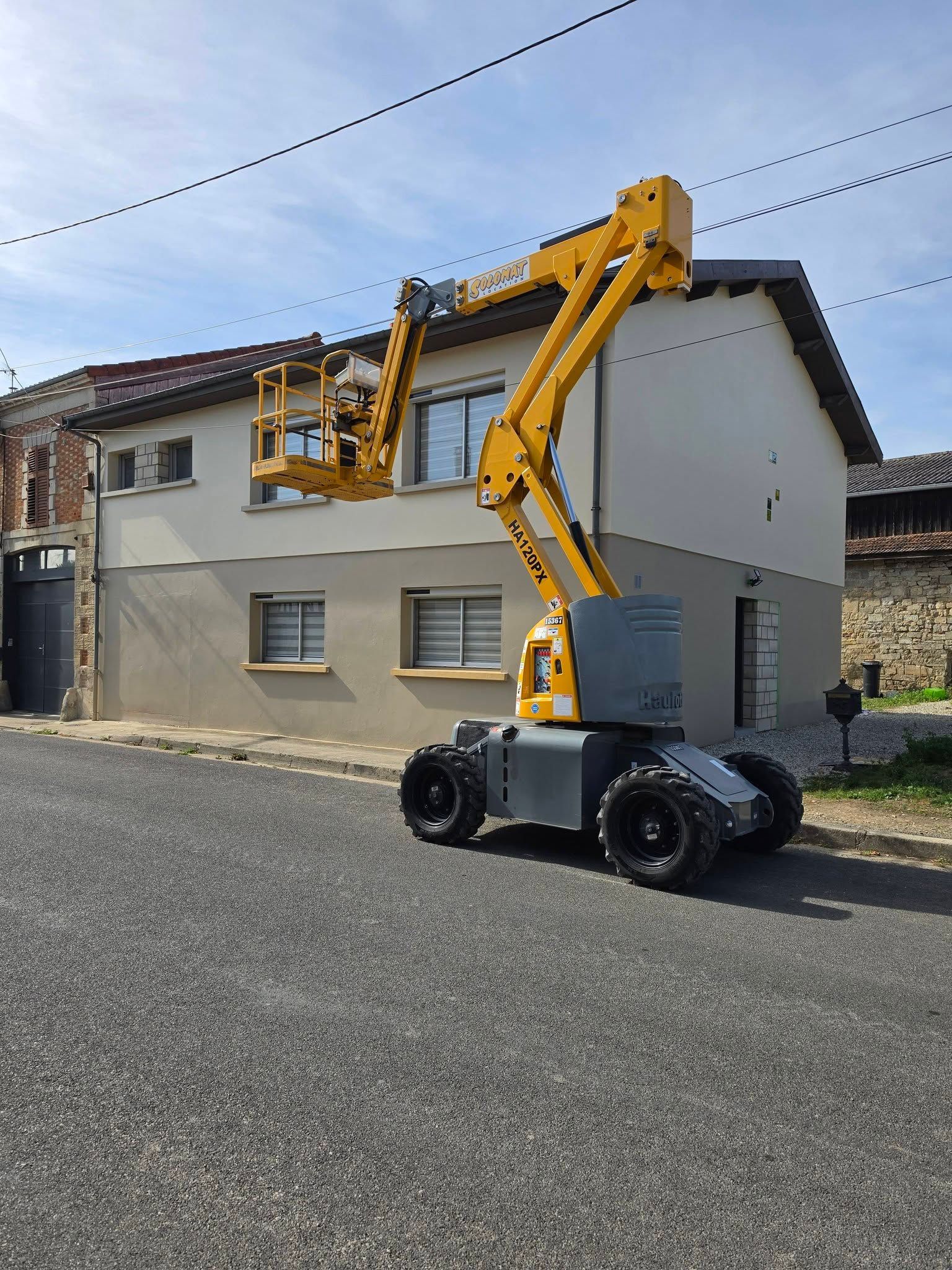 Nacelle élévatrice jaune soulevant une nacelle avec un ouvrier à côté d’une maison beige, dans une rue résidentielle