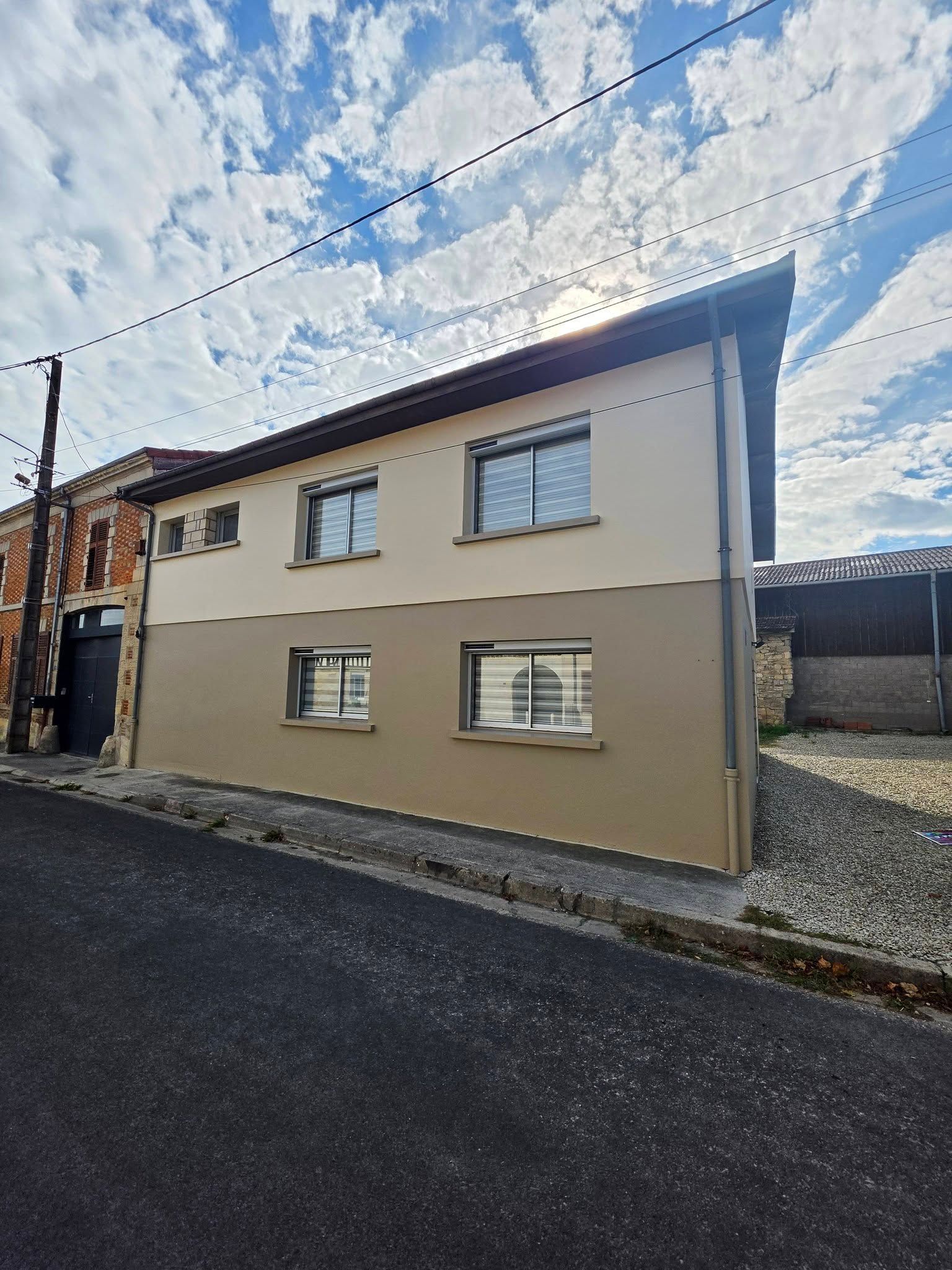 Maison beige à deux étages avec des boiseries blanches, au bord d’une rue étroite sous un ciel nuageux