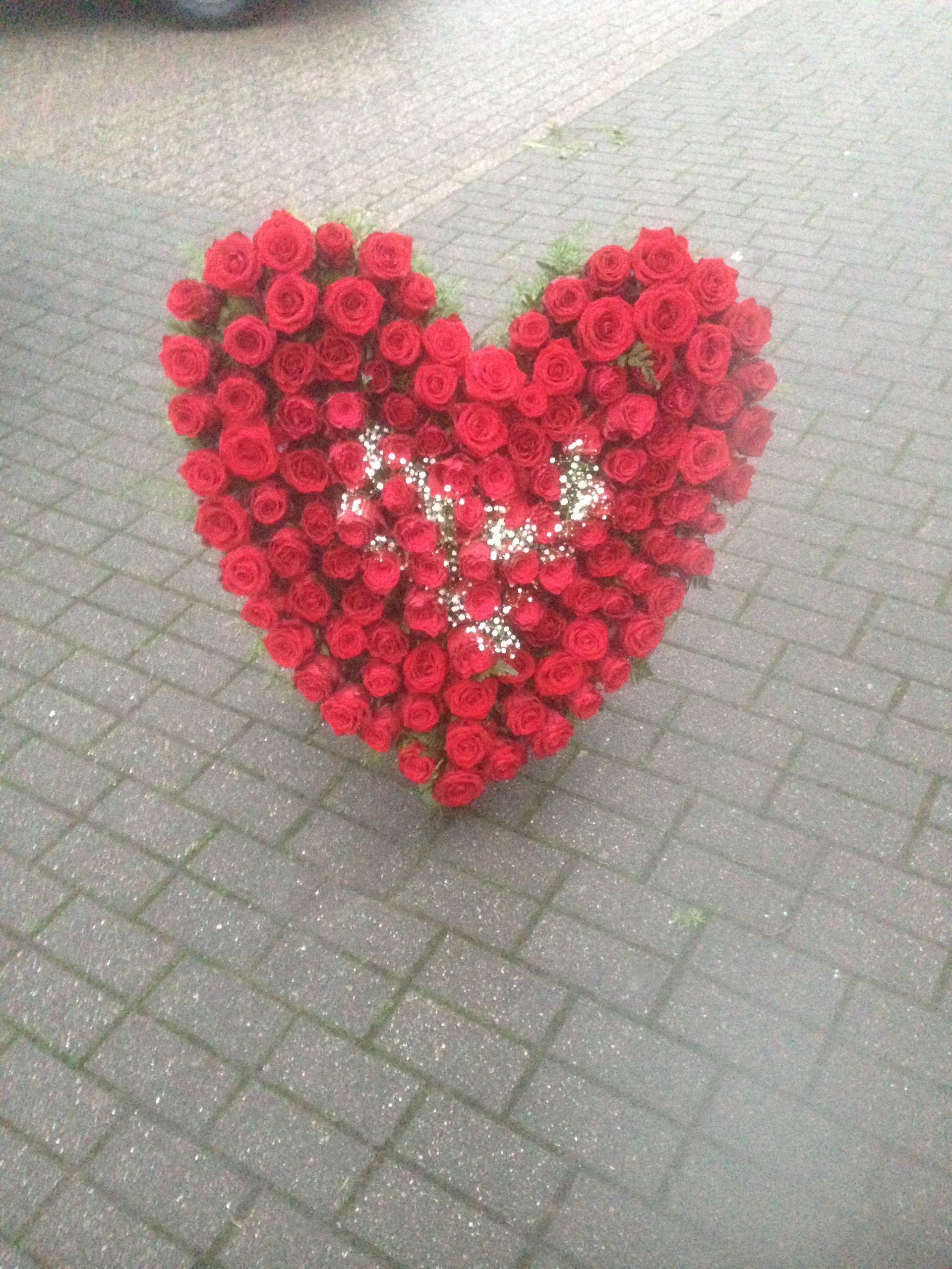 Herzförmiges Arrangement aus roten Rosen auf grauem Backsteingrund, mit einem weißen floralen Element in der Mitte.