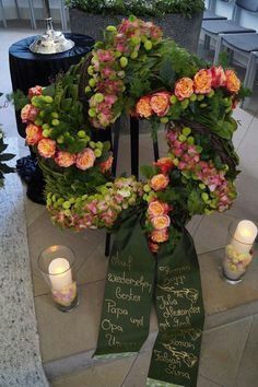 Ein Blumenkranz mit rosa und orangefarbenen Rosen steht auf einem Ständer. Ein grünes Banner mit goldener Schrift ist ebenfalls vorhanden. Zwei Kerzen brennen.
