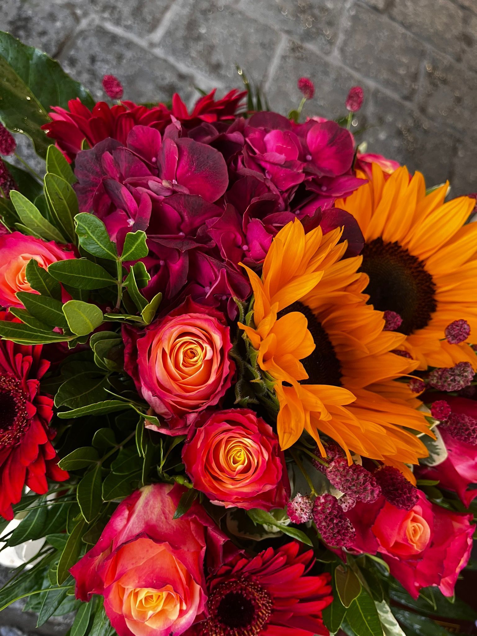 Ein farbenfroher Blumenstrauß mit Sonnenblumen, Rosen und Hortensien in Rot-, Orange- und Dunkelvioletttönen.