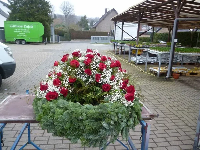 Ein Kranz aus roten Rosen und weißen Blüten auf einem Tisch im Freien vor einem Blumengeschäft, im Hintergrund ein grüner Wohnwagen.