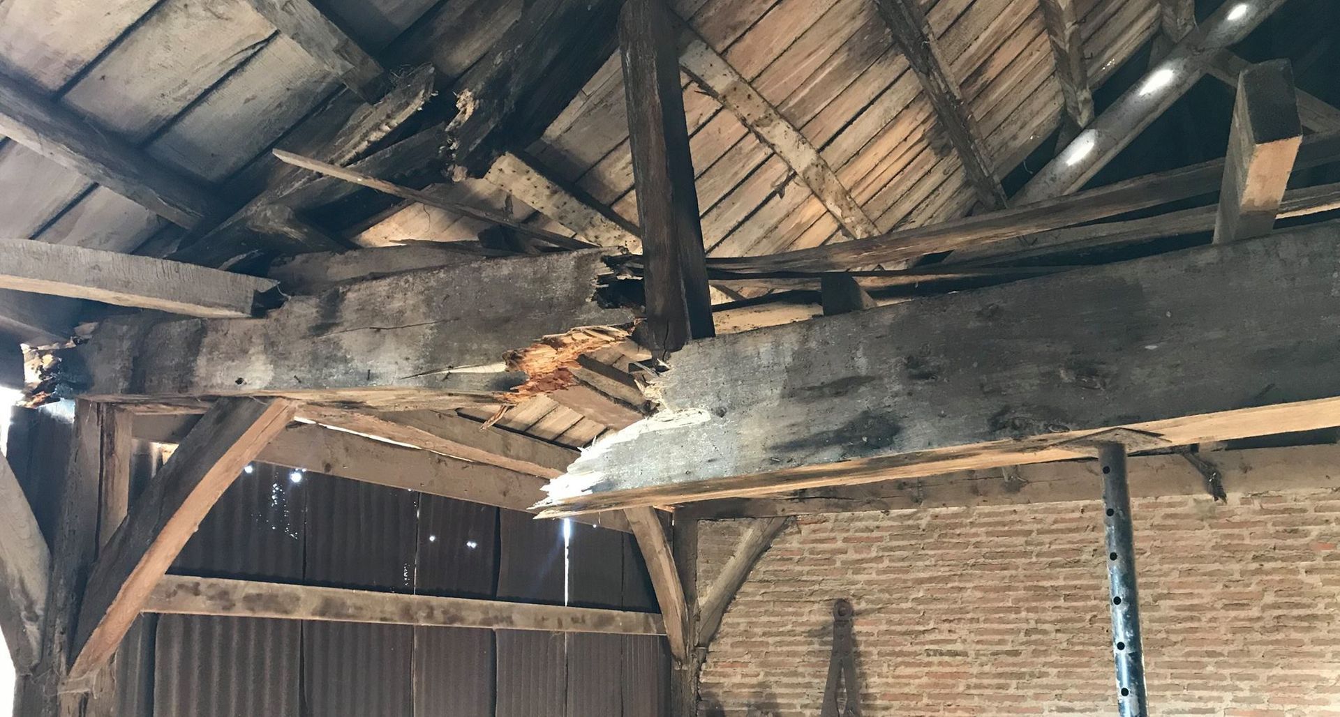 Poutre de grange en bois endommagée, avec des sections qui s'effritent ; la lumière du soleil filtre à travers.