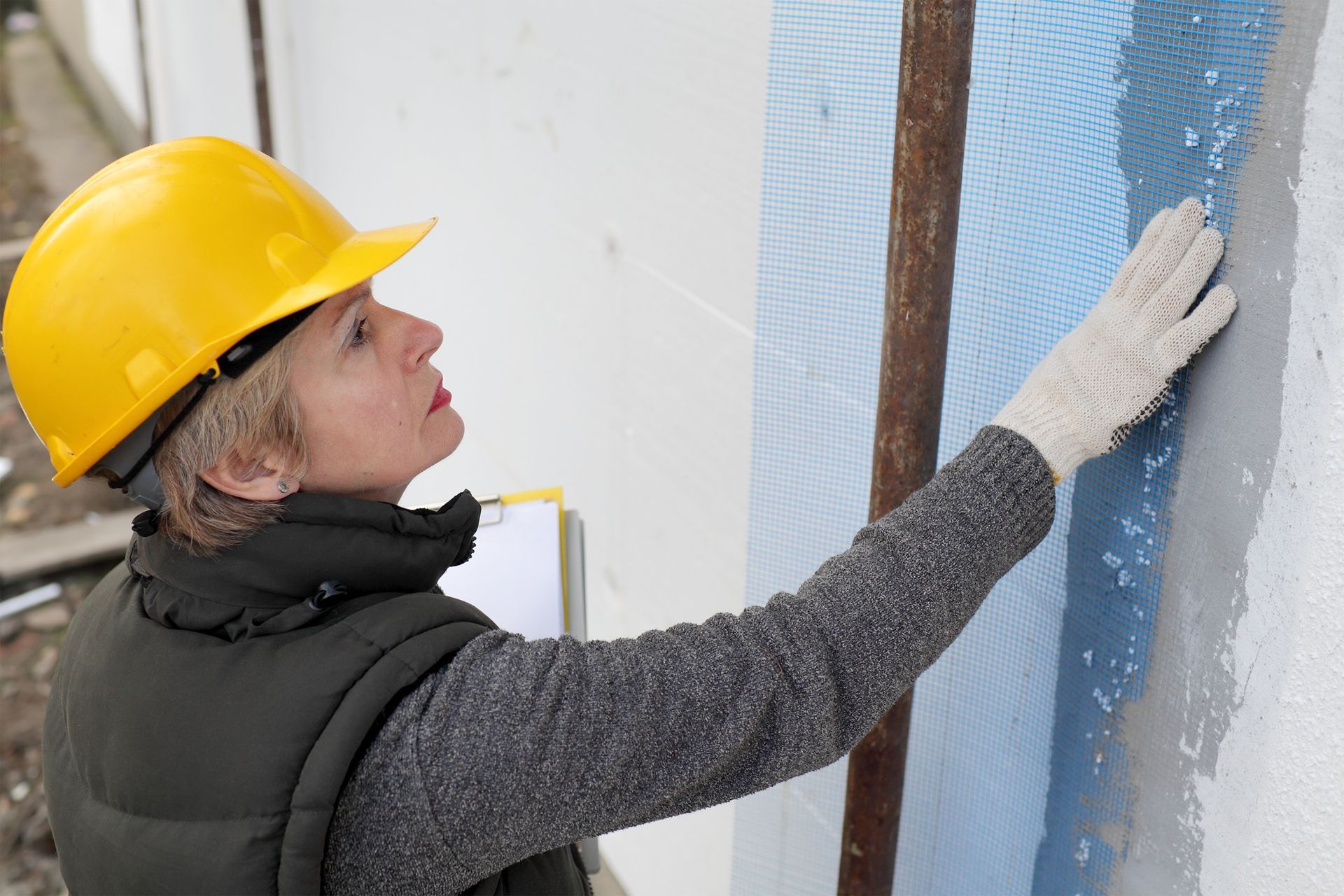 Une personne portant un casque de chantier jaune examine un mur, gantée de travail, à l'extérieur, sur un chantier de construction.