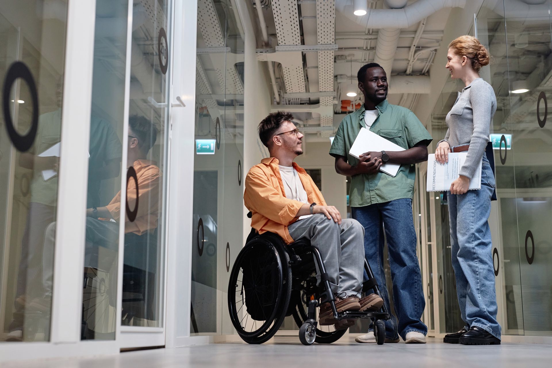 Trois personnes discutent dans le couloir d'un bureau ; une en fauteuil roulant, deux debout avec des documents.
