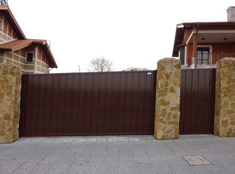 Una puerta corrediza marrón está entre dos pilares de piedra frente a una casa.