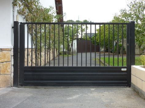 Una puerta negra se abre a un camino de entrada con árboles al fondo.