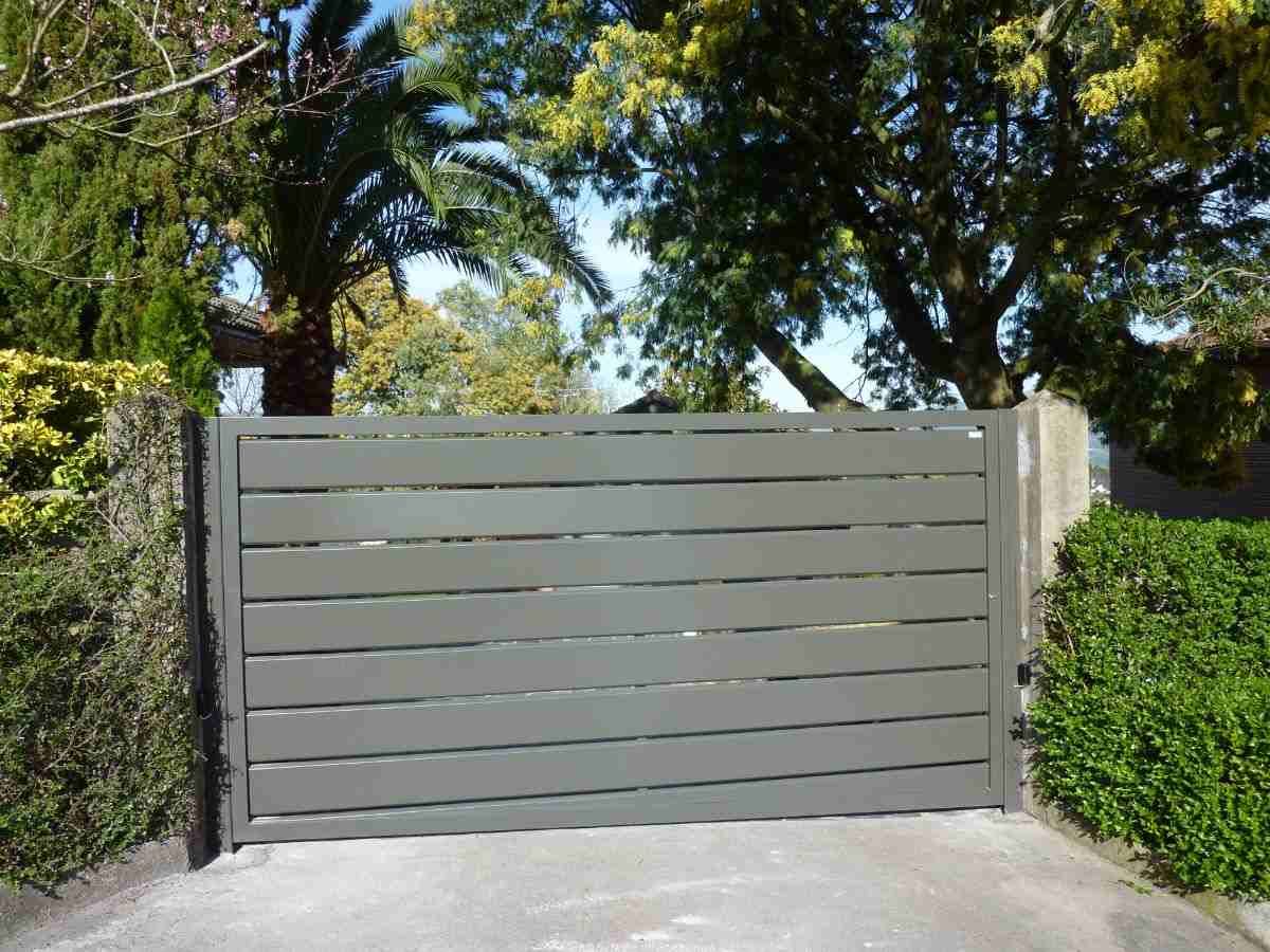 Una puerta gris se abre a un camino de entrada con árboles al fondo.