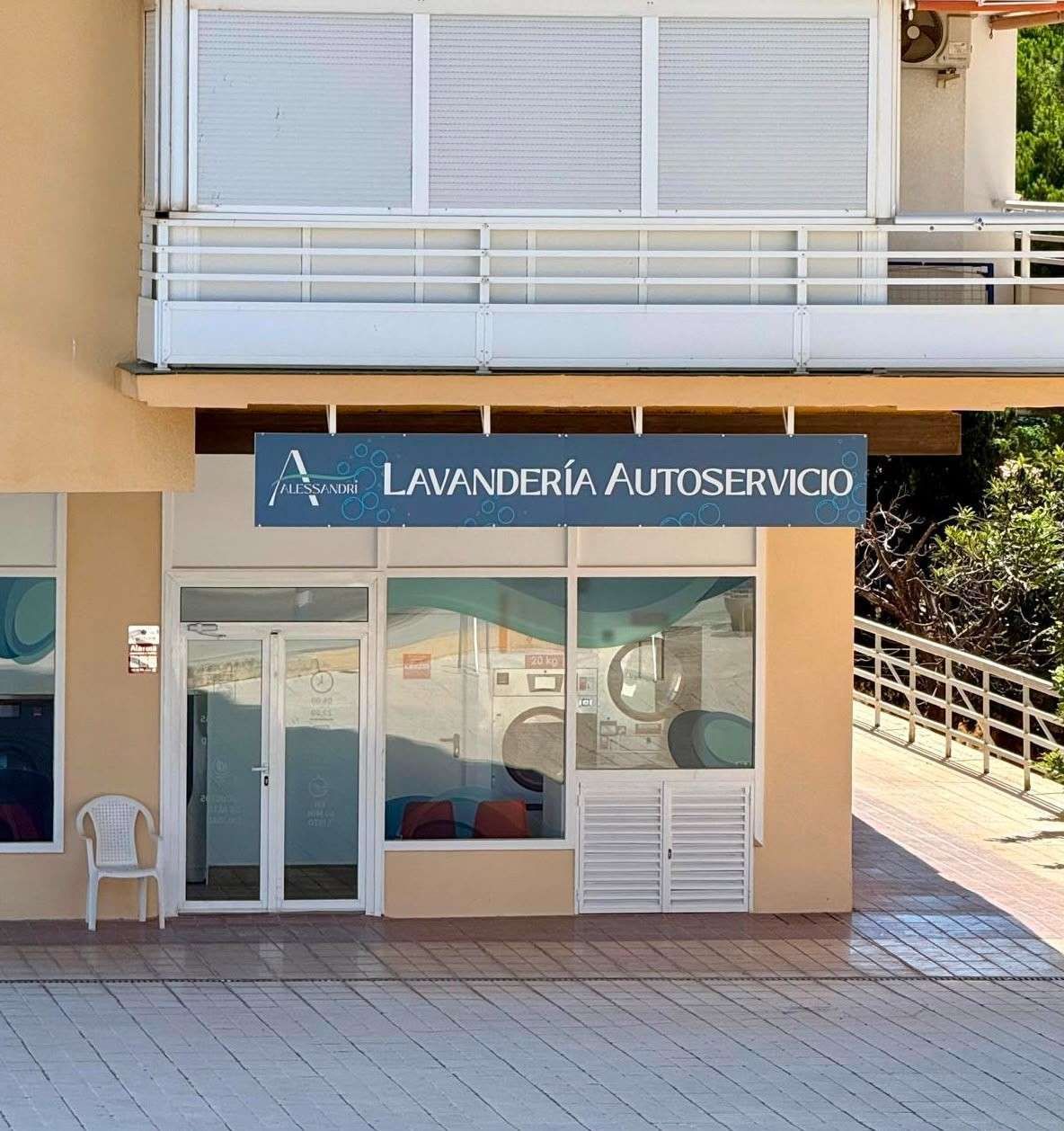 Lavandería de autoservicio con letrero azul y entrada de cristal en la fachada de un edificio soleado.
