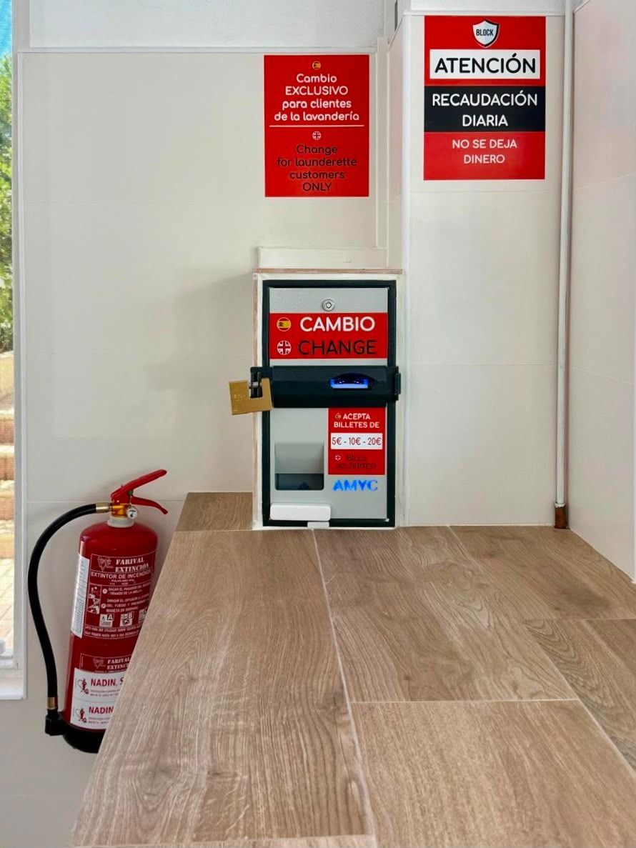 Un extintor de incendios junto a una pared blanca con señales de seguridad rojas y una caja de cambio en el suelo.