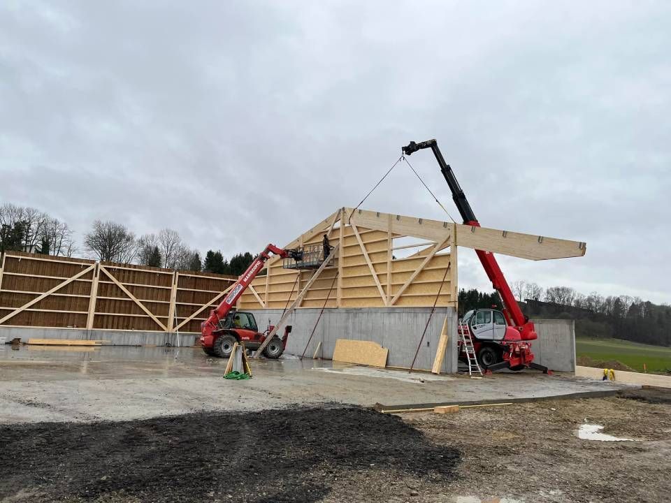 Chantier de construction avec deux chariots élévateurs rouges soulevant une grosse poutre en bois pour la placer sur la charpente d'un bâtiment.