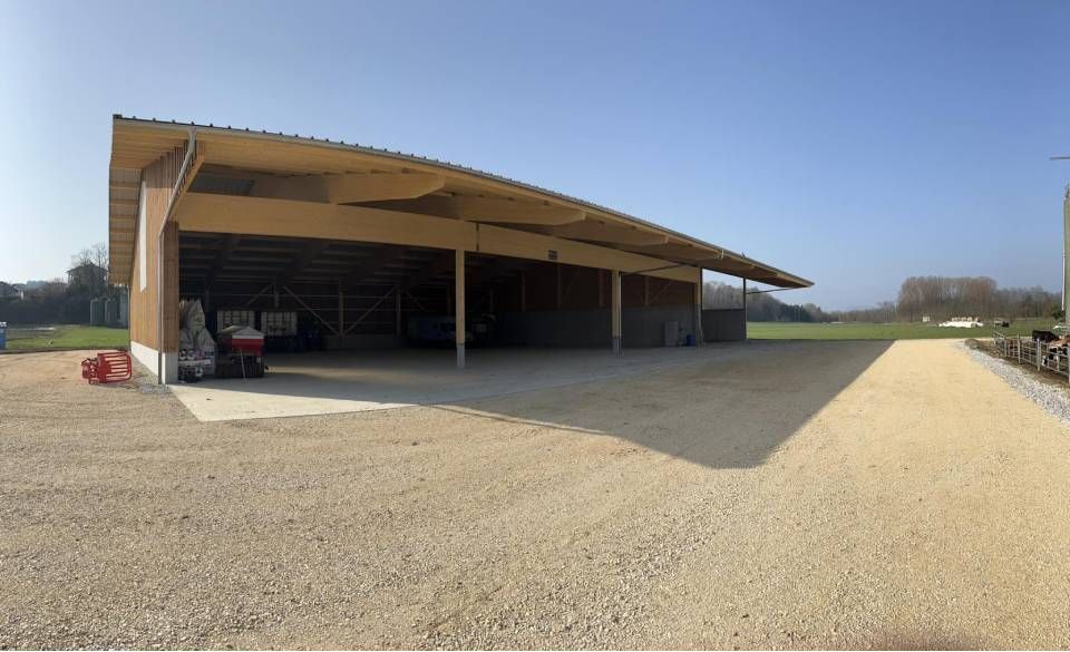 Hangar en bois ouvert sur les côtés, avec parking en gravier, sous un ciel bleu, dans une ferme.