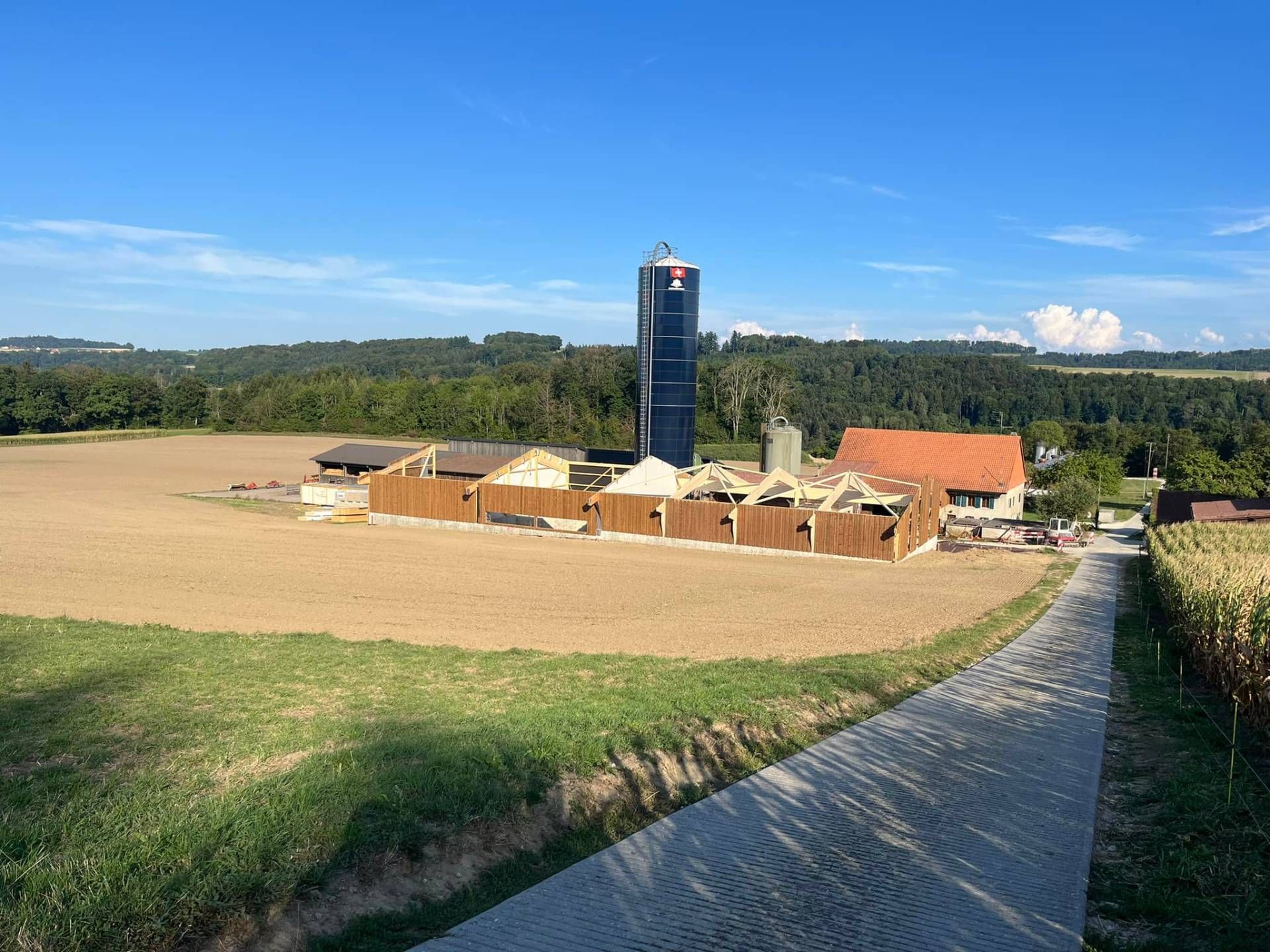 Ferme avec un haut silo, des bâtiments bruns, un champ moissonné et une route sous un ciel bleu.