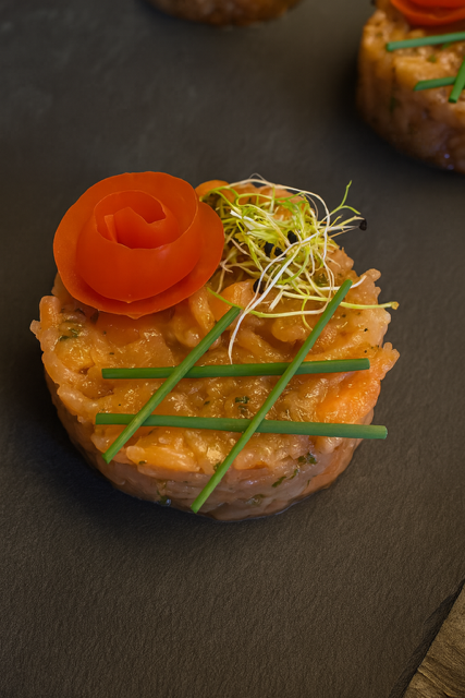 Tartare de saumon, rond, garni de rose de tomate, de pousses et de ciboulette sur une surface foncée.