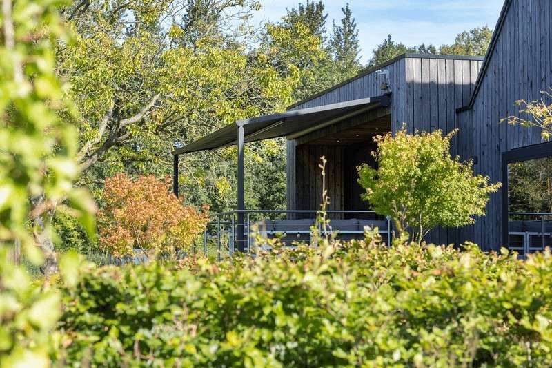 Edificio de madera con techo oscuro y porche, rodeado de exuberante follaje verde y naranja.