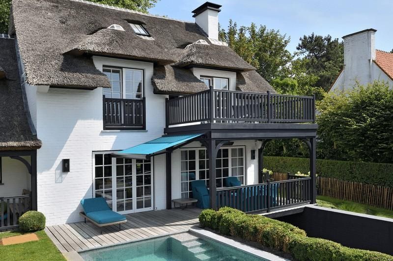 Casa blanca con techo de paja, balcón, toldo azul y piscina.