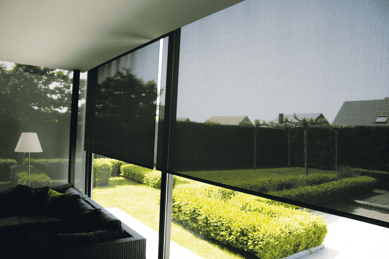 Persianas enrollables negras que cubren grandes ventanales con vistas a un jardín verde con setos y césped.