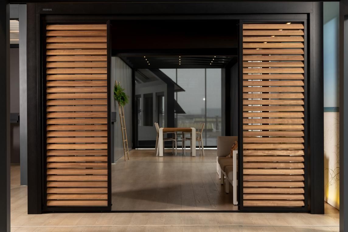 Un toldo negro cubre los asientos al aire libre de un edificio con grandes ventanales. Un coche circula por la calle.