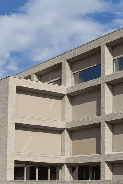 Edificio de ladrillo beige con ventanas en forma de cuadrícula y un cielo azul brillante con nubes blancas.
