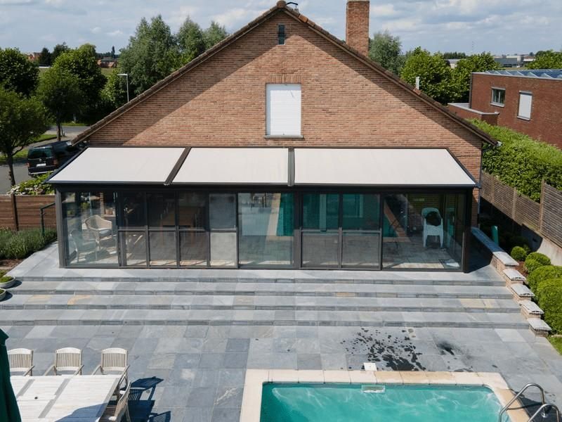 Terraza acristalada adosada a una casa de ladrillo, sobre un patio de piedra gris con piscina.