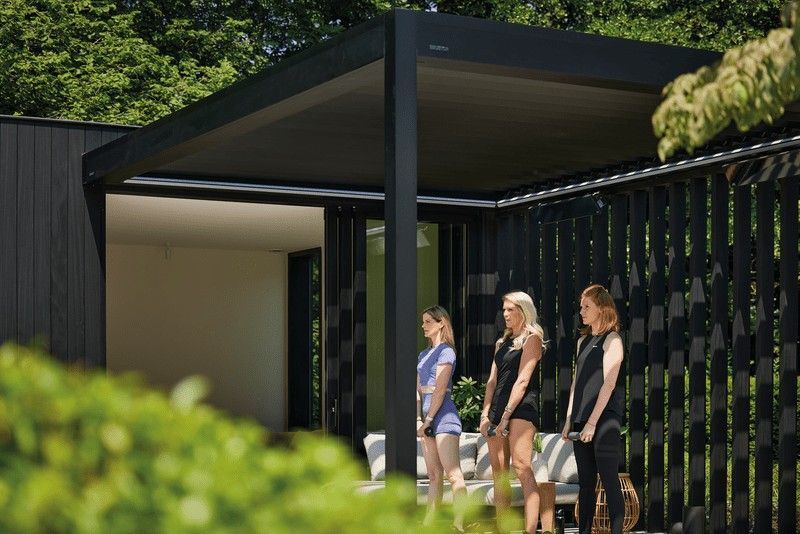 Tres mujeres de pie bajo una pérgola moderna junto a una valla de listones.