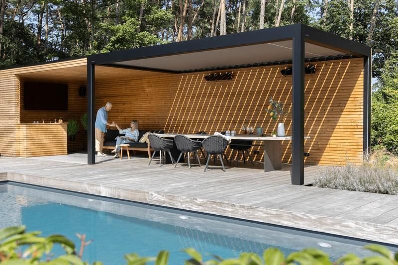 Comedor al aire libre con piscina. Una pérgola cubierta da sombra a una mesa con asientos. La gente socializa.