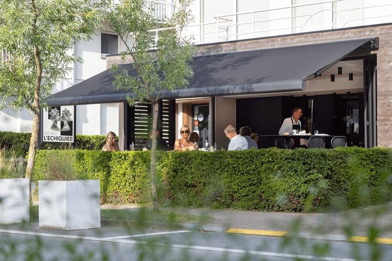 Patio del restaurante con toldo negro, seto verde y gente sentada en mesas.