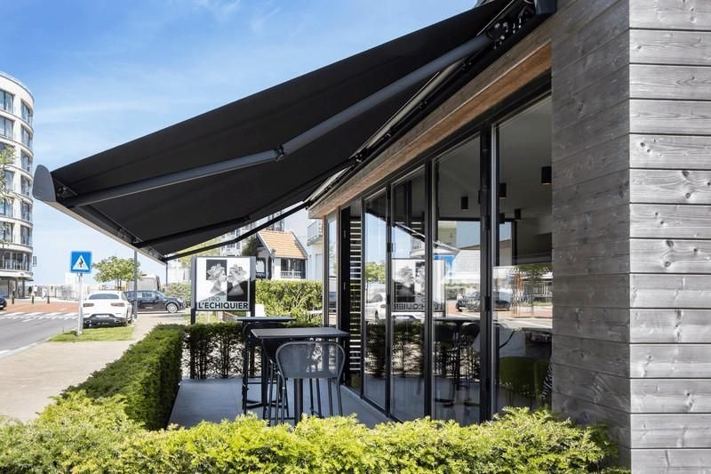Un toldo negro cubre los asientos al aire libre de un edificio con grandes ventanales. Un coche circula por la calle.