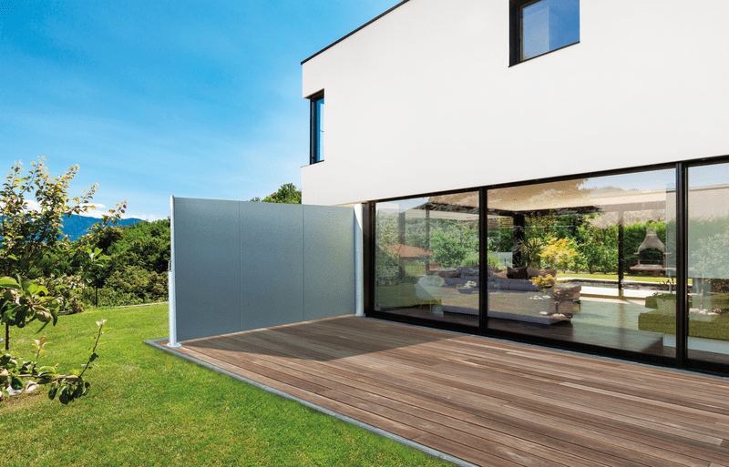 Casa moderna con mampara de privacidad gris sobre una terraza de madera con vistas a un jardín con montañas.