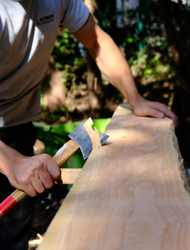 Une personne utilise une hache pour façonner une longue planche de bois à l'extérieur.