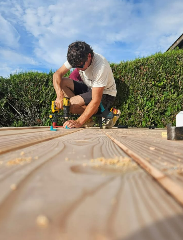 Une personne utilise une perceuse pour construire une terrasse en bois à l'extérieur.
