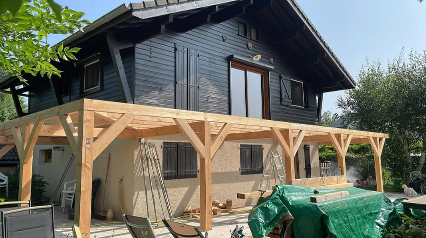 Porche en bois en construction sur une maison sombre, avec des supports en bois et des matériaux de construction devant.
