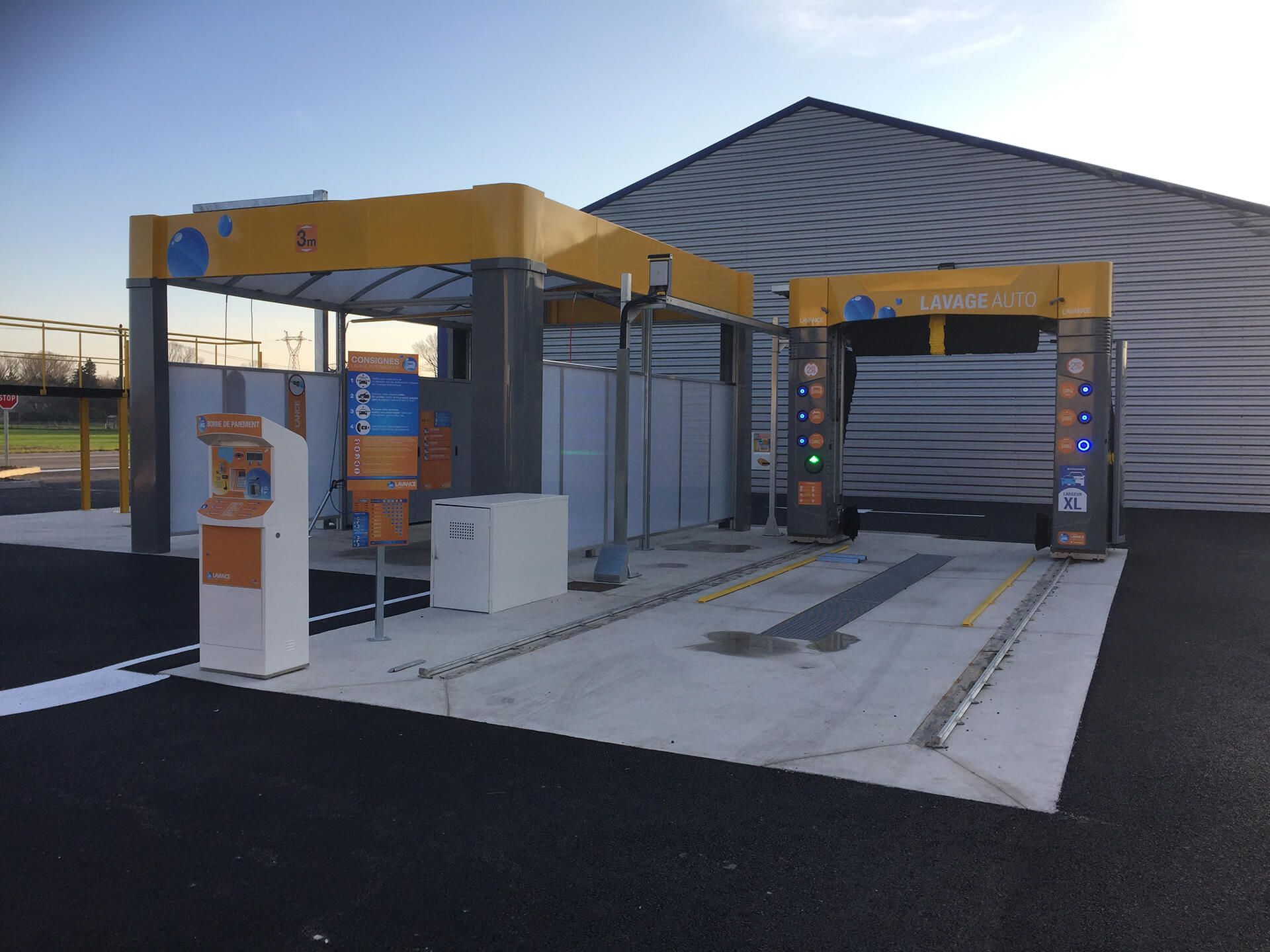 Station de lavage de voitures avec des structures jaunes et grises, comprenant une borne de paiement et des baies de lavage.