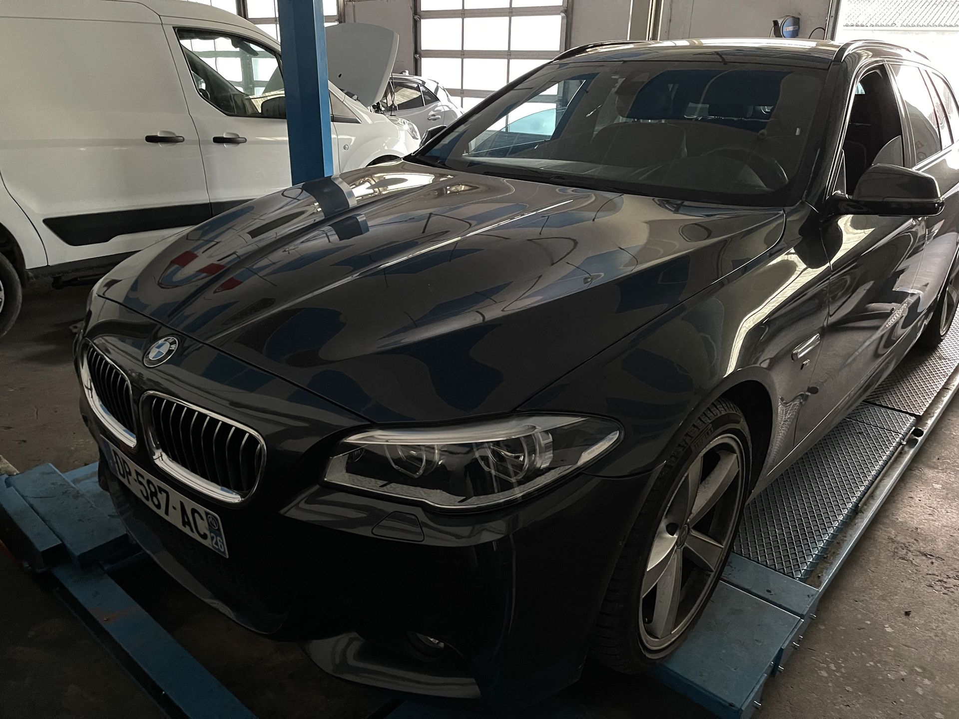 Un break BMW noir dans un garage, sur un pont élévateur, avec une camionnette blanche en arrière-plan.