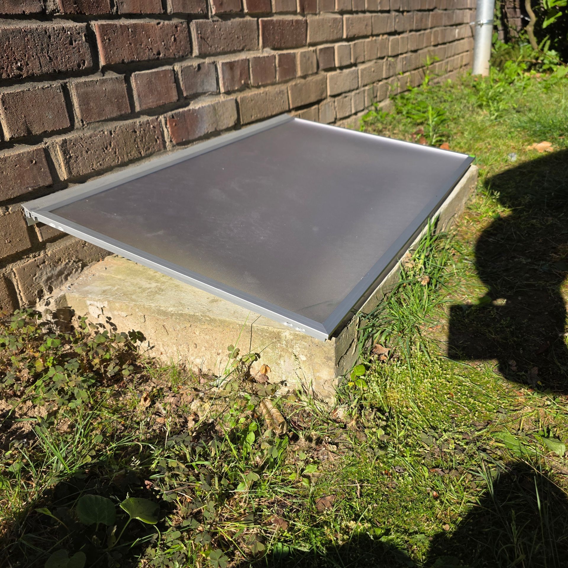 Graue Metallkellertür neben einer Backsteinmauer, eingefasst in einen Betonrahmen über Gras und Unkraut.