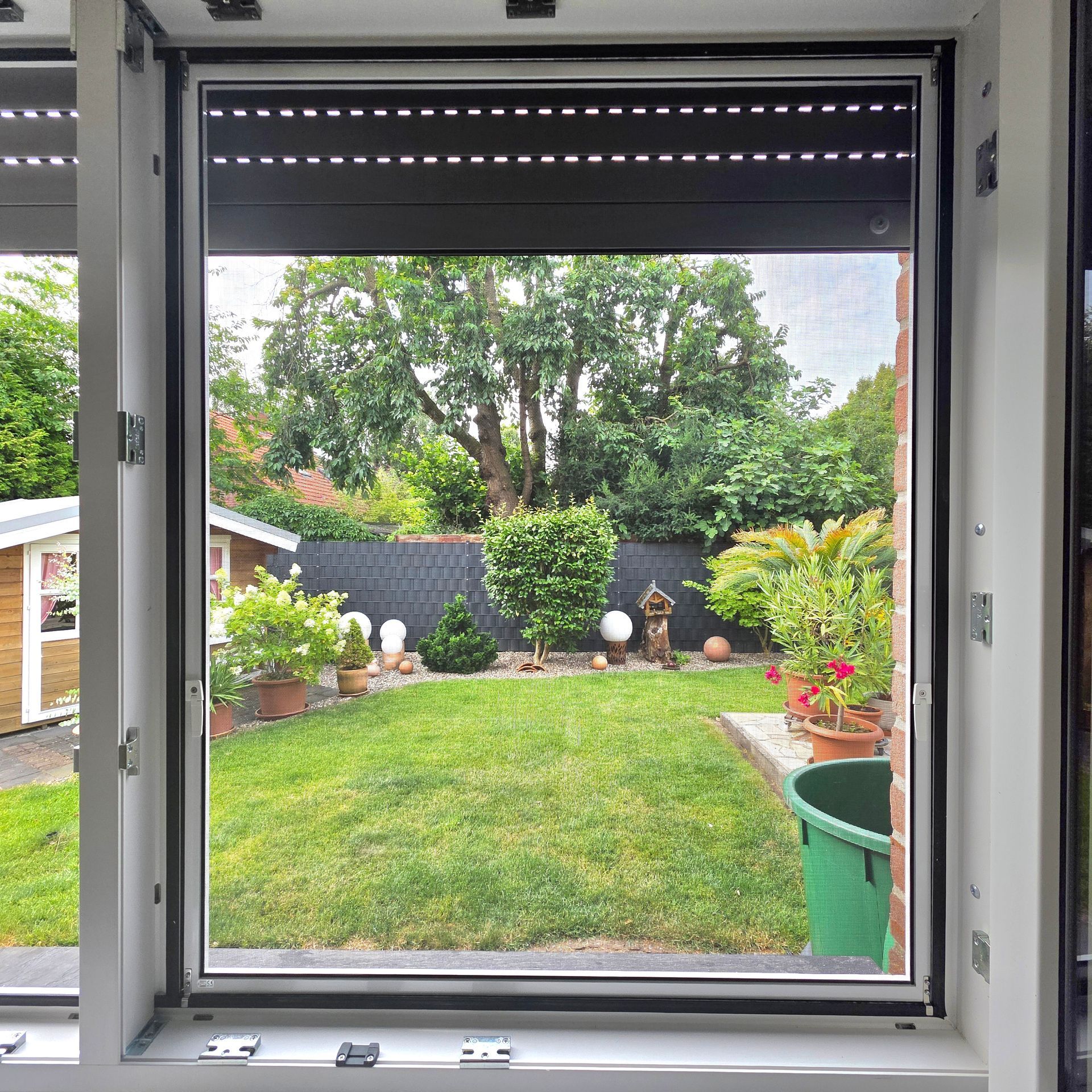 Blick aus einem Fenster auf einen grünen Hinterhof mit Rasen, Sträuchern, Bäumen und Terrassendekoration.