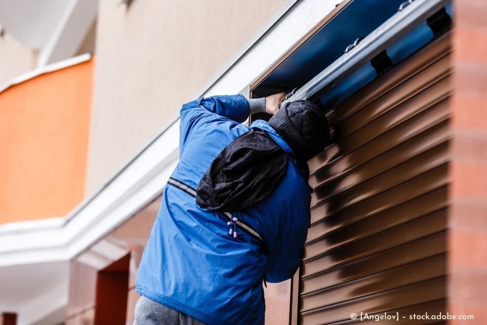 Eine Person in einer blauen Jacke öffnet einen braunen Metallrollladen an der Fassade eines Gebäudes.