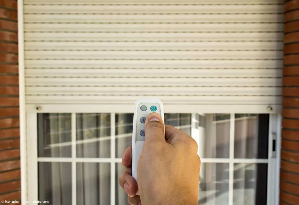 Eine Hand hält eine Fernbedienung, die auf einen weißen Fensterladen über einem Fenster an einer Backsteinwand gerichtet ist.