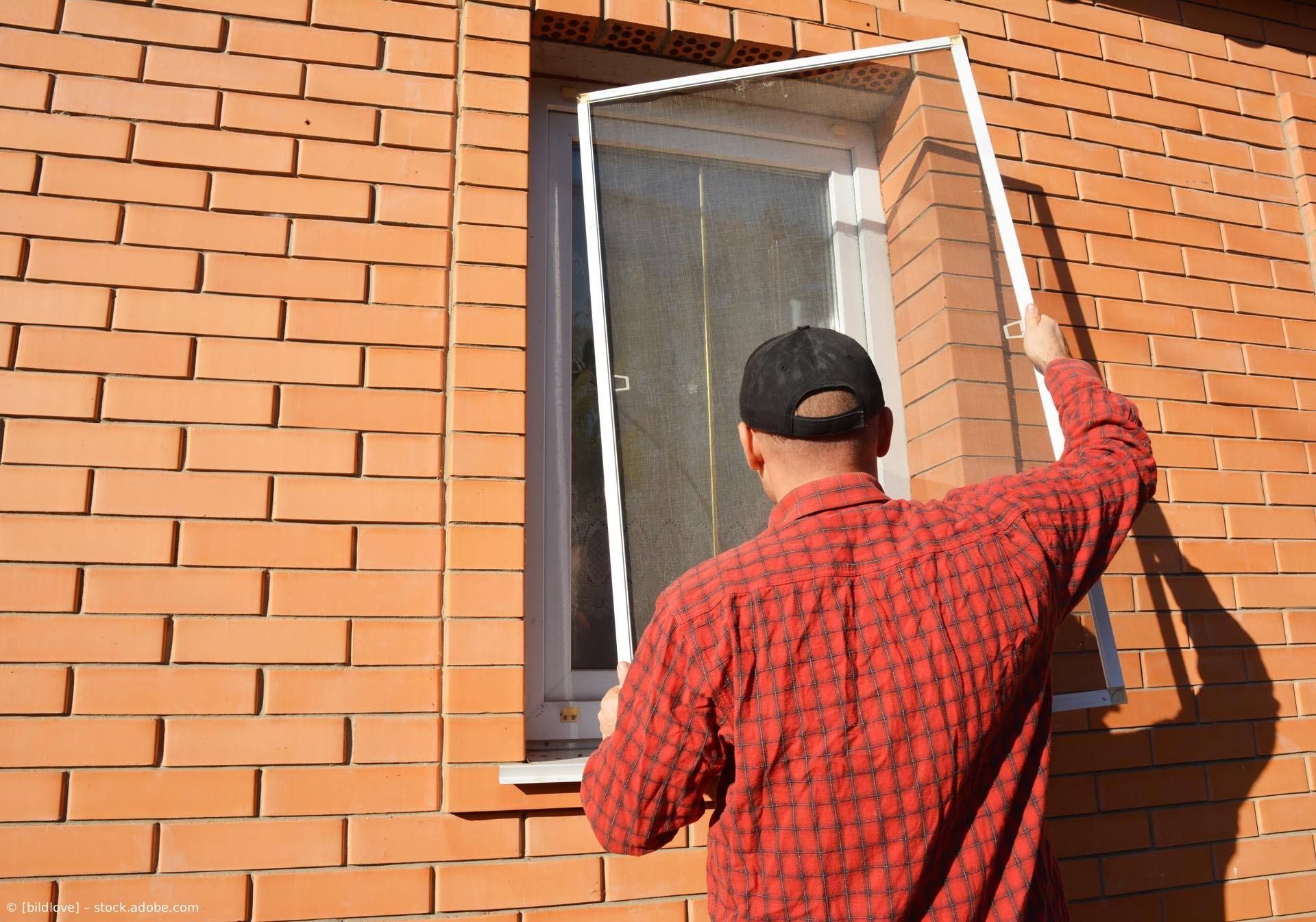 Eine Person in einem rotkarierten Hemd montiert einen weißen Fensterrahmen an einer Backsteinmauer.
