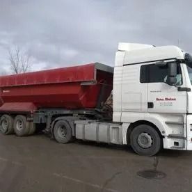 Un camión semirremolque blanco arrastrando un remolque basculante rojo por una carretera mojada bajo un cielo nublado.