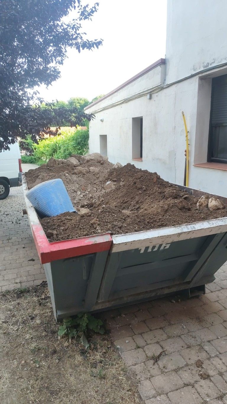 Un gran contenedor de basura lleno de tierra se encuentra junto a un edificio blanco, cubierto con una lona azul.