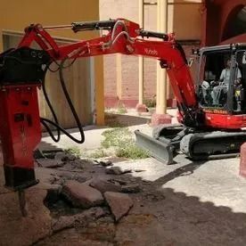 Una excavadora roja rompe el hormigón en un patio o entrada de vehículos.