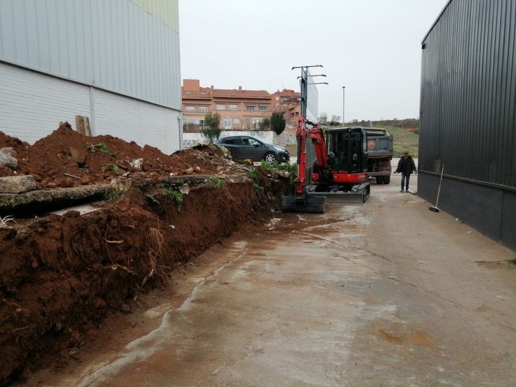 Un estrecho camino embarrado con una excavadora, montones de tierra y dos personas cerca de edificios industriales.