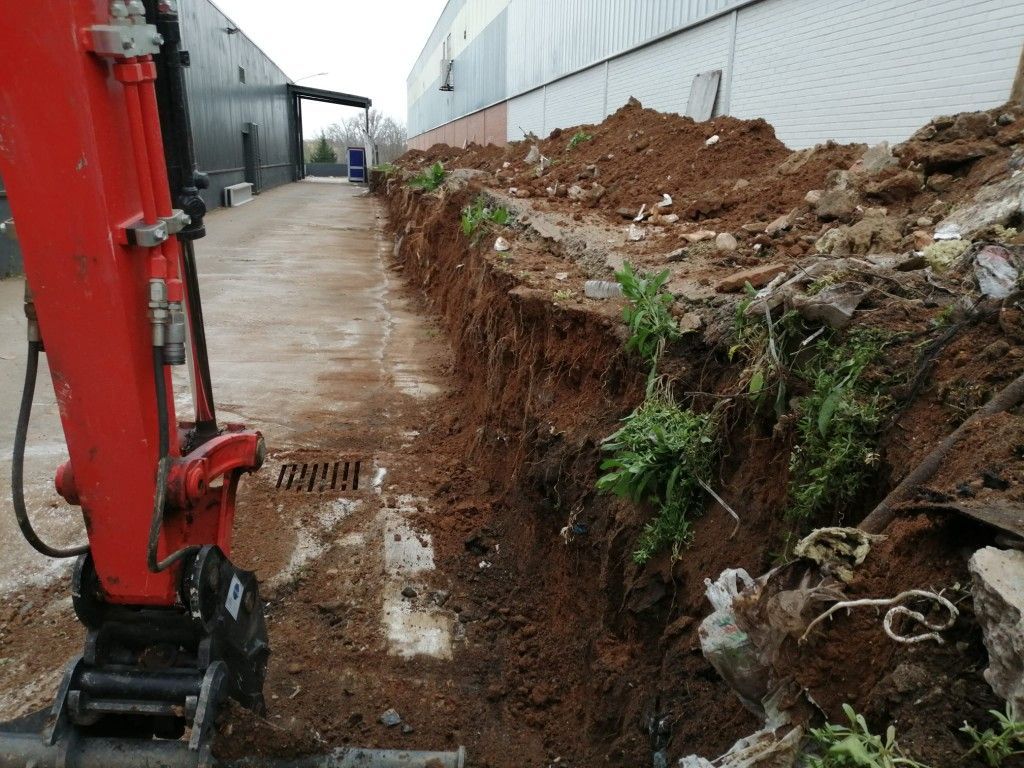 Excavadora roja junto a una zanja fangosa al lado de un edificio y un montón de tierra en un callejón industrial.