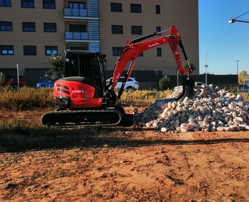 Excavadora roja excavando en un montón de rocas en una obra en construcción cerca de un edificio.