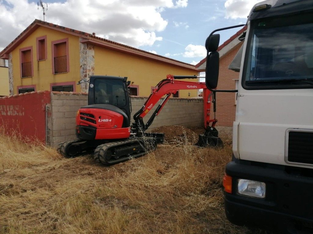 Una miniexcavadora roja junto a un camión blanco en un patio seco al lado de una casa color canela.