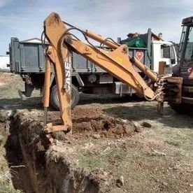 Una excavadora amarilla cava una zanja junto a un camión en una obra de construcción.